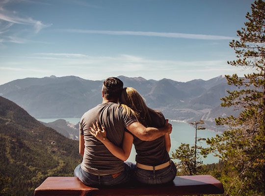 A man and a woman sitting on a bench and looking out at a mountain range